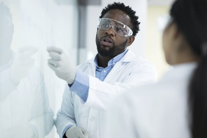 Photo shows a scientist speaking with colleagues at a white board in a laboratory/Getty Images