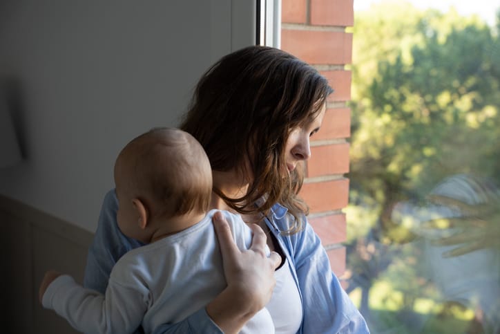 Photo shows a new mother holding her baby while looking out the window (Getty Images/Rolart Studio)
