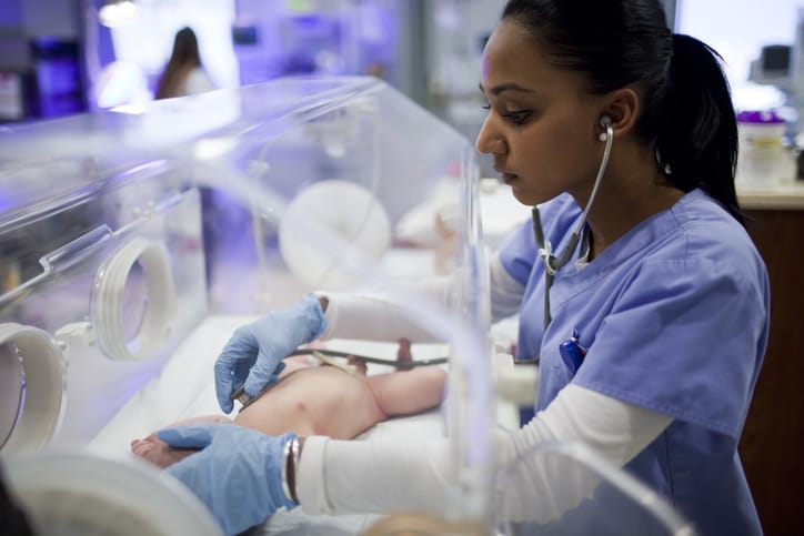Photo shows a nurse listening to a newborn's heartbeat in an incubator/Getty Images/RubberBall Productions