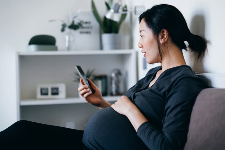 Photo shows a pregnant woman sitting on a couch on her phone at home (Getty Images/d3sign)